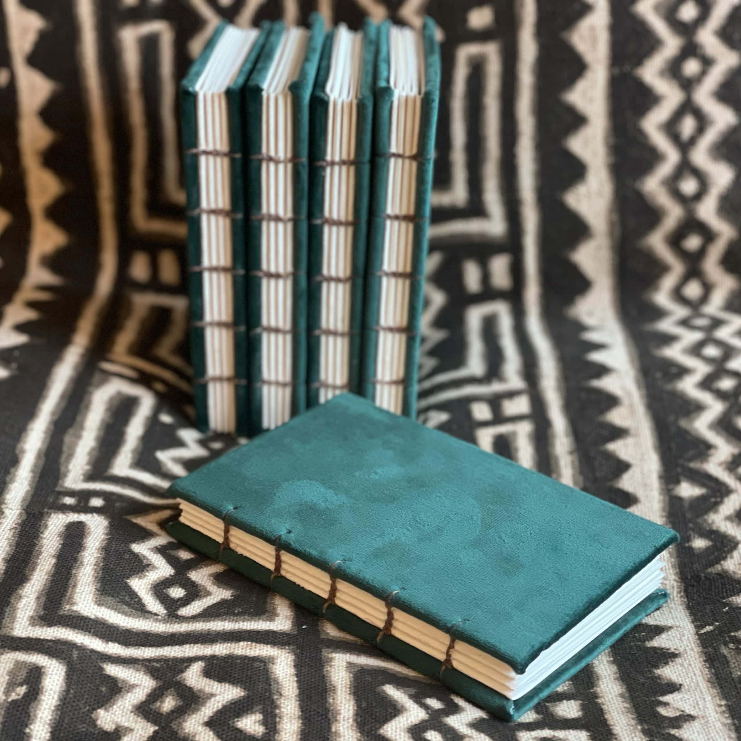 This is an image of handmade sketchbooks. This particular image features four of them standing upright against an African patterned blanket, with one laying down in front of the others. All four books feature a clear image of the binding